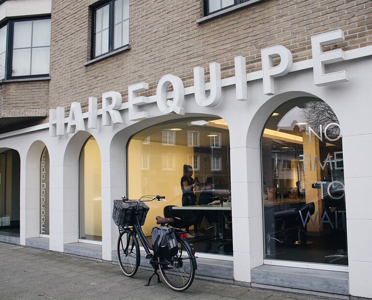 Gevel van een kapsalon met de naam "HAIREQUIPE" en een fiets met mand voor de deur. Een persoon is binnen zichtbaar.