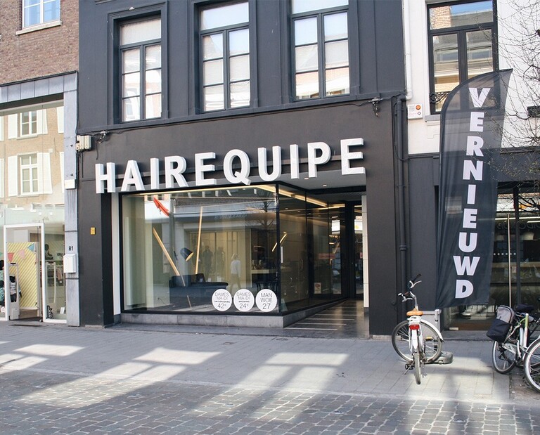 Gevel van een kapsalon genaamd "HAIREQUIPE", met ramen en een vlag met het woord "VERNIEUWD", fietsen staan geparkeerd voor de deur.