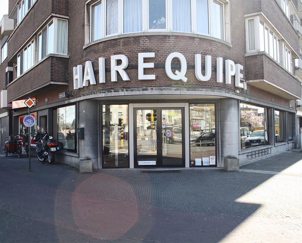 Gevel van een kapsalon met de naam "HAIREQUIPE", zichtbaar met grote letters, en een ingang met glazen deuren; motorfietsen en fietsenparkeren in de omgeving.