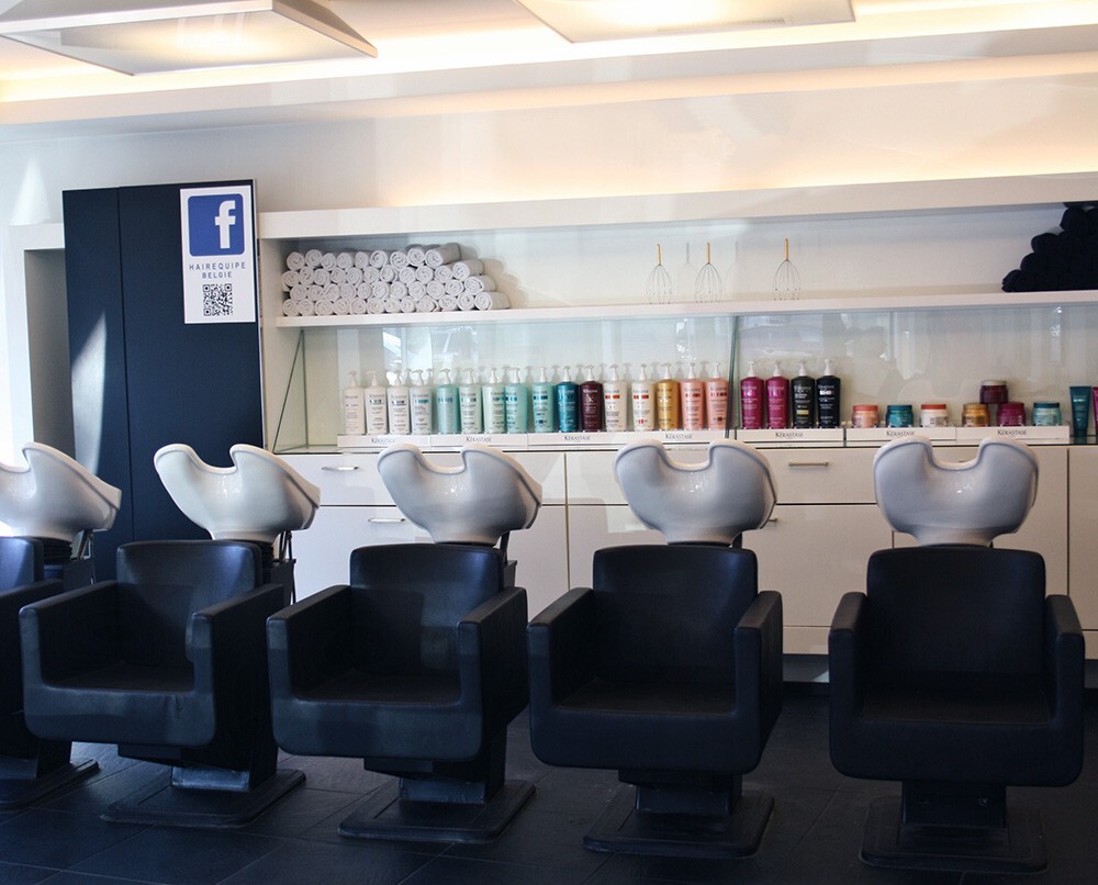 Interieur van een kapsalon met drie wasstoelen, een muur met verzorgingsproducten en gestapelde handdoeken.