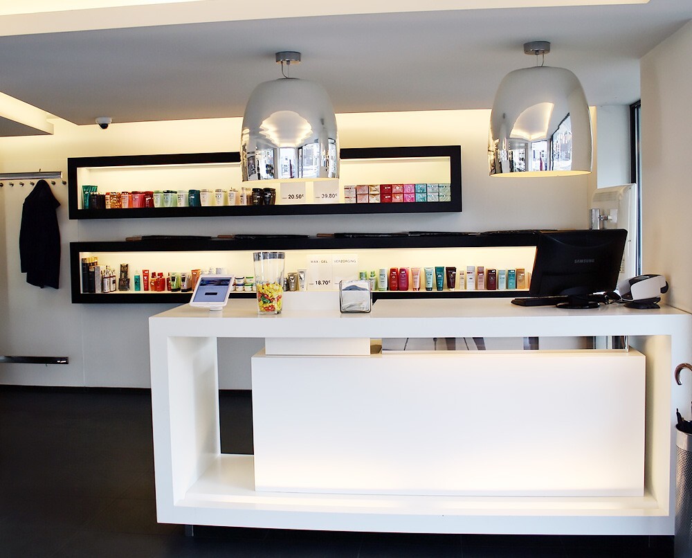 Een moderne receptie met een witte balie, glazen schalen met snoep, een tablet en een computerscherm, omgeven door kleurrijke producten op planken boven de balie. Twee opvallende hanglampen verlichten de ruimte.