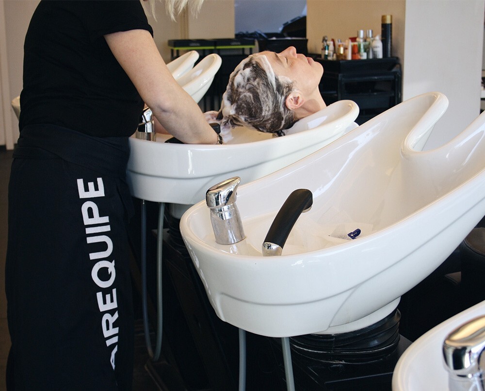 Een haarsalon waarin een stylist het haar van een klant wast in een wasbak, met enkele andere lege wassen in de achtergrond.