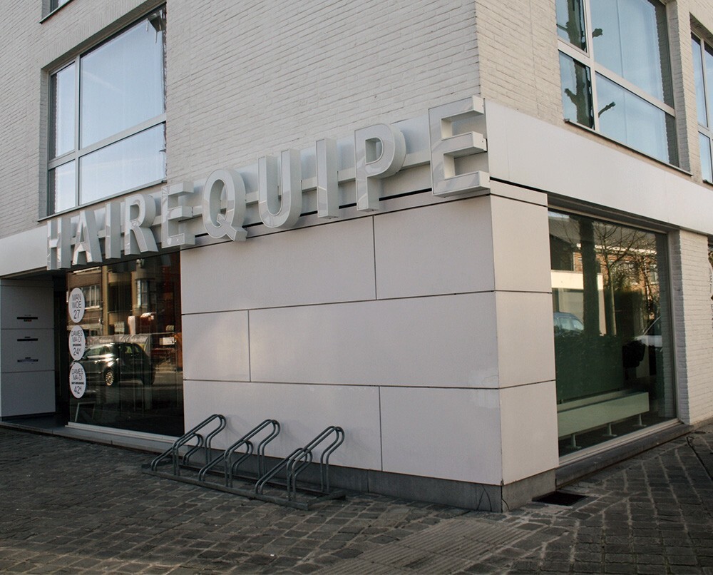 Een modern gebouw met de naam "HAIREQUIPE" in groot, verlicht letters aan de onderste hoek, met een glazen gevel en fietsenstalling.