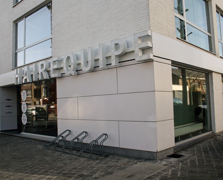 Een modern gebouw met de naam "HAIREQUIPE" in groot, verlicht letters aan de onderste hoek, met een glazen gevel en fietsenstalling.
