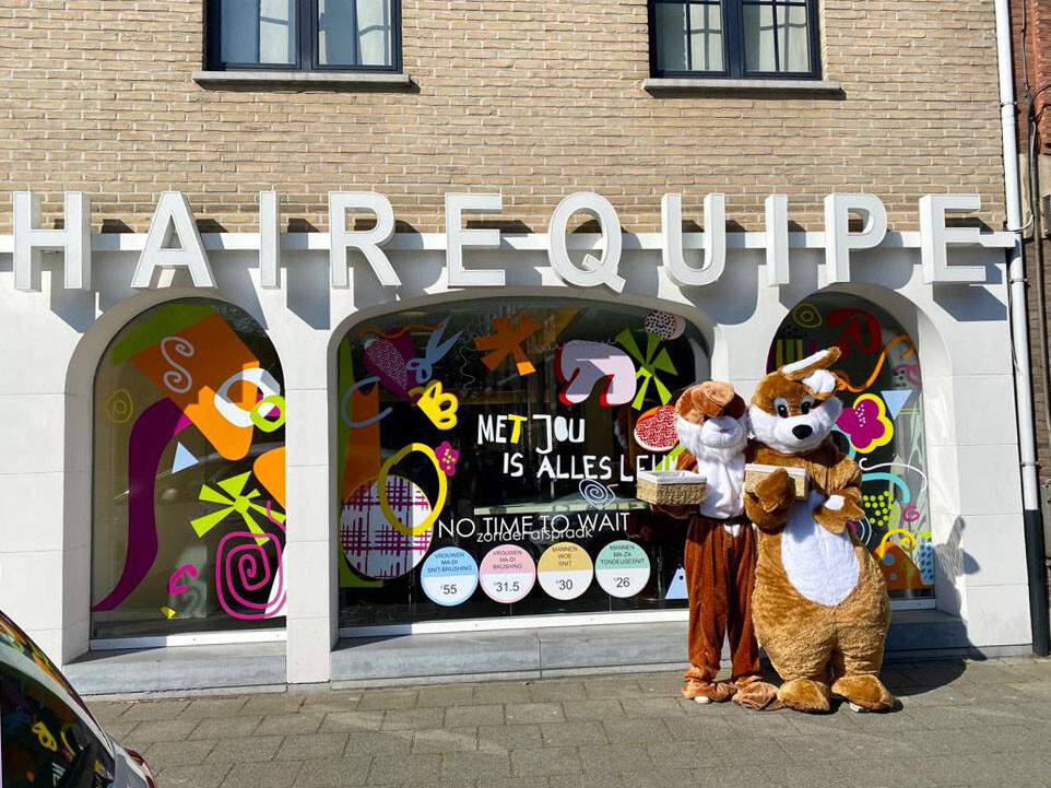Twee mensen in dierenkostuums staan voor een kapsalon met kleurrijke ramen en de tekst "MET JOU IS ALLES LEUK".