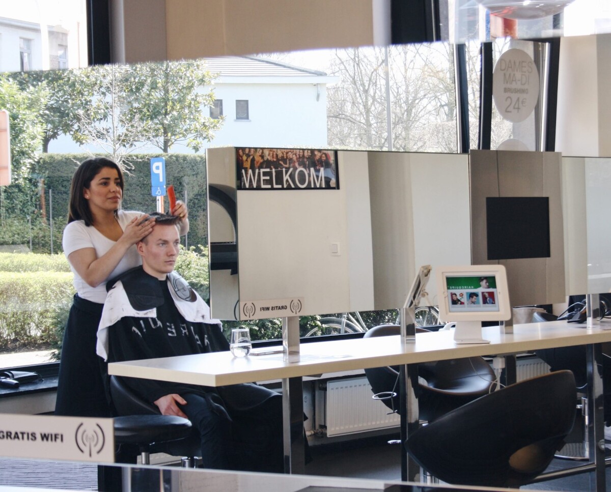 Vrouw kapselt een man met een schort in een moderne kapsalon, terwijl een spiegel verschillende reflecties toont.