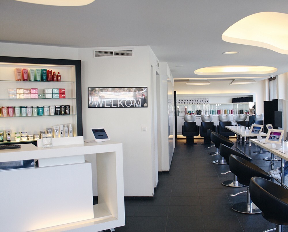 Interieur van een moderne salon met een receptiebalie, producten op een schap, en stoelen voor klanten. Een bord met de tekst &quot;WELKOM&quot; is zichtbaar.