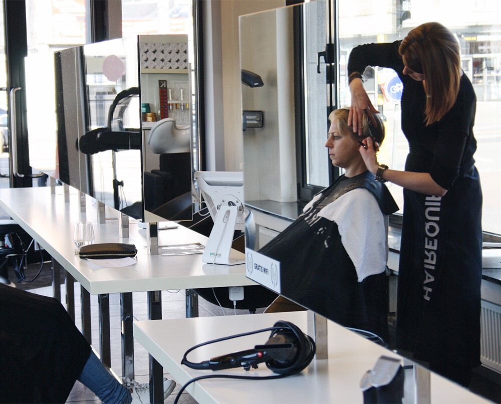 Een kapster knipt het haar van een klant in een moderne kapsalon met spiegels en een lichte, minimalistische inrichting.