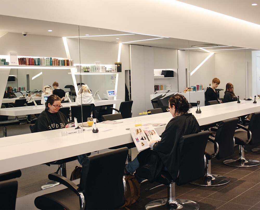 Een moderne kapsalon met meerdere klanten die in spiegels kijken, terwijl ze tijdschriften lezen of op hun telefoons zitten. Aan de wand zijn verzorgingsproducten en een wastafel zichtbaar.