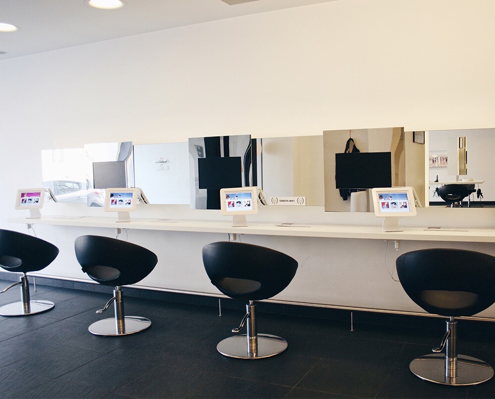 Interieur van een moderne salon met een lange witte tafel, spiegels en zwarte stoelen, elk voorzien van een tablet.