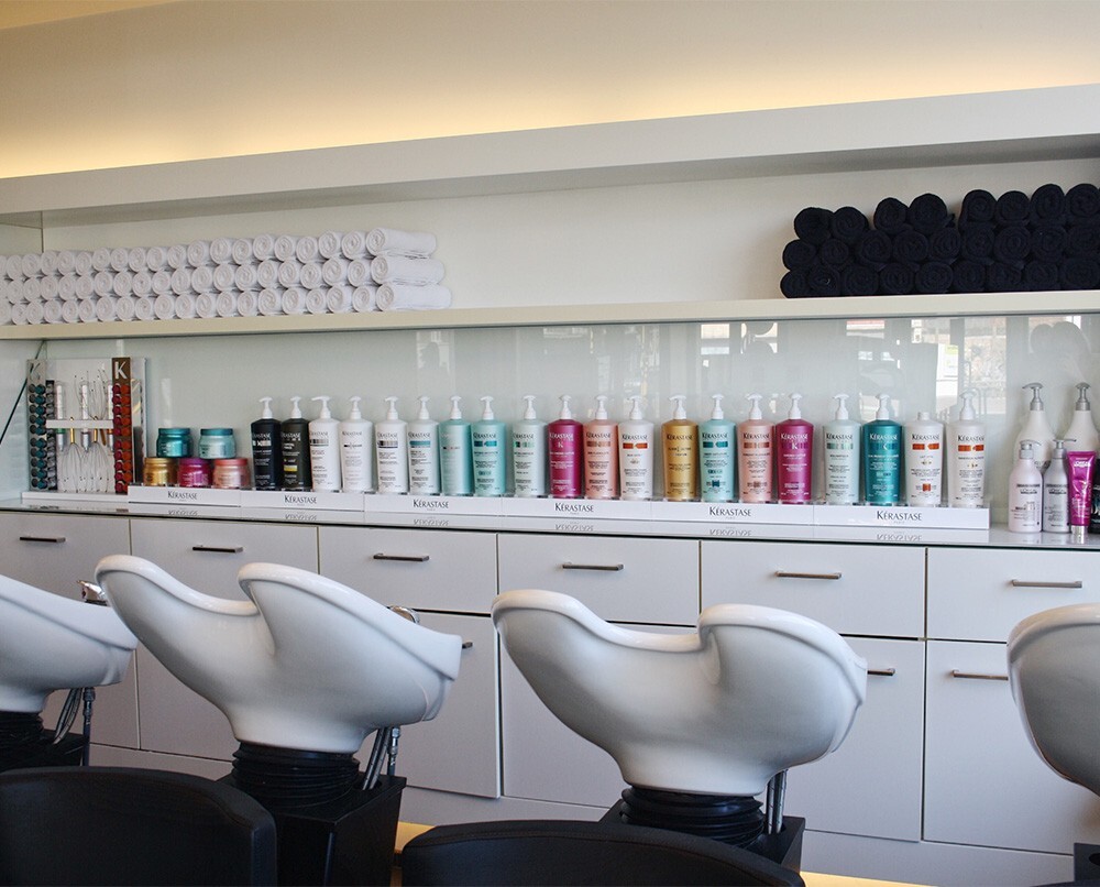 Interieur van een kapsalon met een rij shampoo- en verzorgingsproducten op een plank, naast opgerolde handdoeken en haarwasstations.