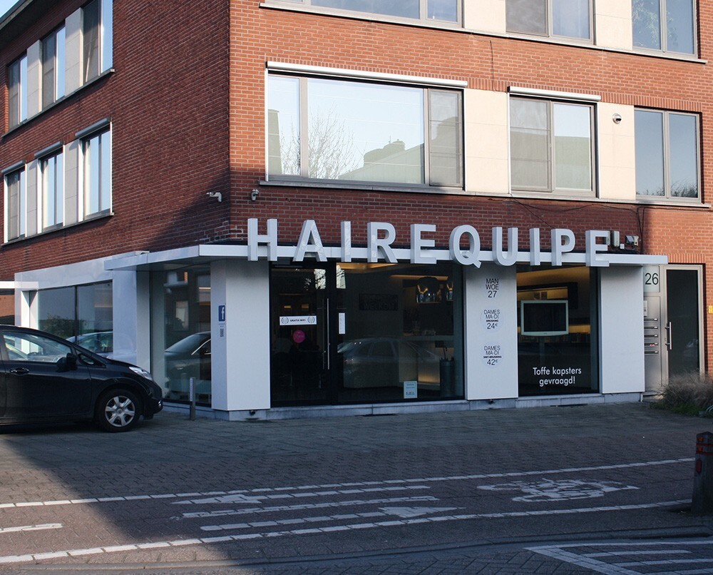 Voorgevel van een haarsalon genaamd &quot;HAIREQUIPE&quot;, met grote letters boven de ingang en informatie over openingstijden en prijzen op een bord.