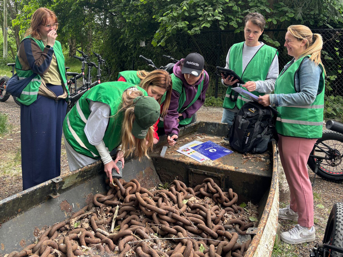 Groep mensen in groene vesten kijkt naar een boot vol met slangen, terwijl ze verschillende materialen en een tablet bespreken. Op de achtergrond zijn fietsen zichtbaar.