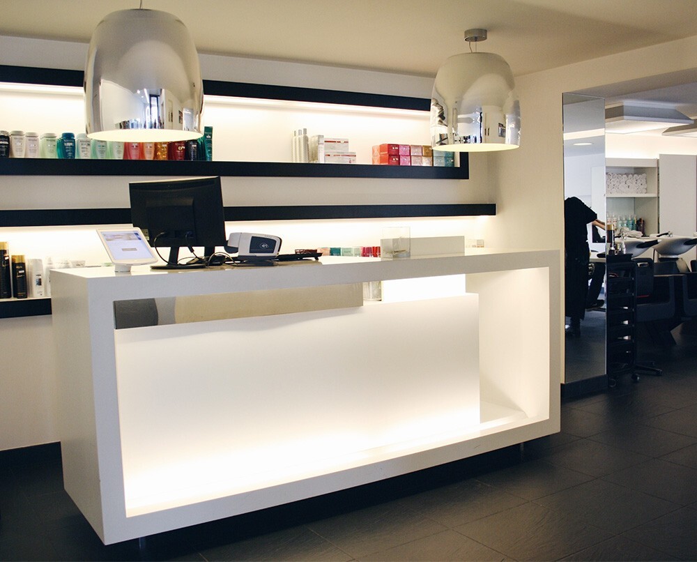 Ingang van een moderne salon met een witte balie, een computer, en producten op planken boven de balie, verlicht door een stijlvolle hanglamp.