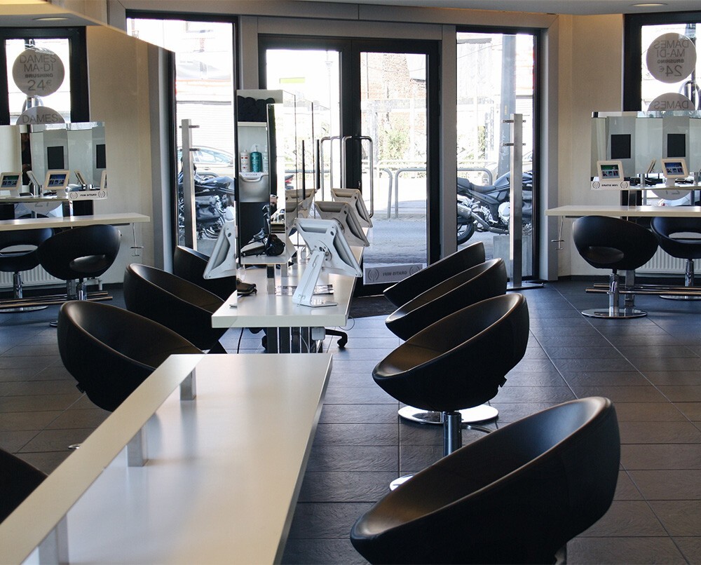 Interieur van een moderne kapsalon met zwarte, ronde stoelen en witte tafels, met spiegels en elektronische apparaten voor klanten.