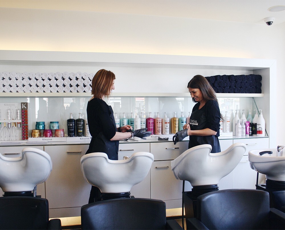 Twee kapsters in een salon bespreken producten, omringd door haarverzorgingsflessen en opgerolde handdoeken, terwijl achter hen een rij wasbakken zichtbaar is.