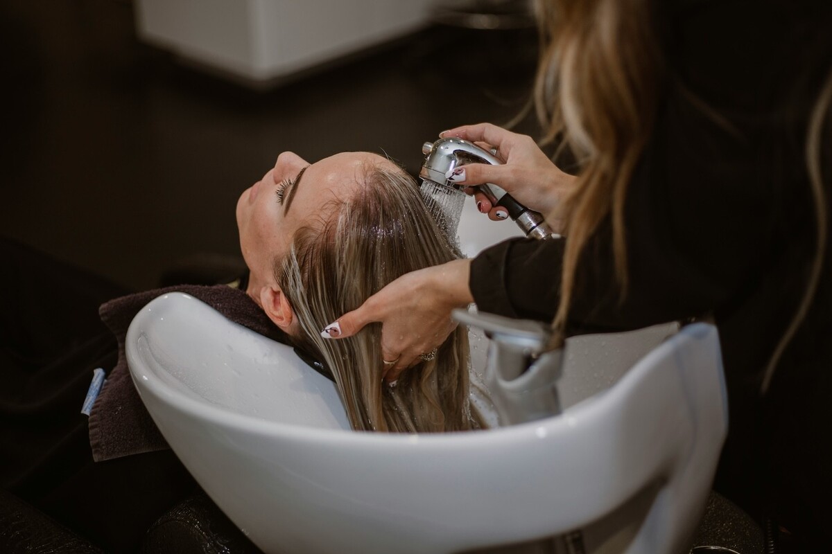 Een vrouw krijgt haar haar gewassen in een kapsalon, terwijl een styliste water over haar hoofd sproeit.